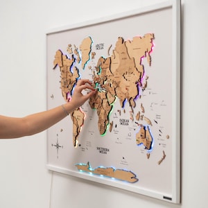 May include: Wooden world map in a white frame, with countries cut out and illuminated by colourful LED lights. A hand points to a location on the map. The map is a decorative item.