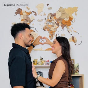 Puede incluir: Un mapa del mundo de madera en tonos marrones y beige est&aacute; montado en una pared blanca. El mapa est&aacute; etiquetado como "M prime Multicolor". Una pareja est&aacute; frente al mapa, formando un coraz&oacute;n con sus manos alrededor de un peque&ntilde;o avi&oacute;n.