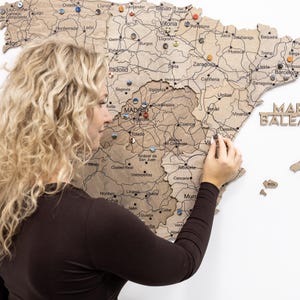 Peut inclure: Une carte en bois de l'Espagne avec les noms des villes et les routes grav&eacute;s. Des &eacute;pingles color&eacute;es marquent les lieux. Une personne place une &eacute;pingle sur la carte. La carte est fix&eacute;e sur un mur blanc.