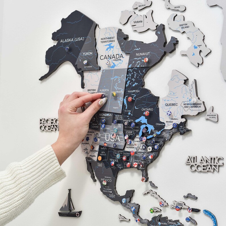 May include: A multi-layered wooden wall map of North America featuring a color palette of black, grey, and white. The map displays country, state, and province names with various small circular flag pins inserted into specific geographic locations.