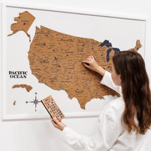 Peut inclure: Une carte en liège encadrée des États-Unis avec les noms des États. La carte est marron avec des épingles bleues et colorées. Une personne place une épingle sur la carte. Une rose des vents et les mots "Pacific Ocean" sont également visibles.
