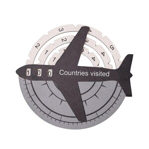 May include: A black airplane-shaped tracker with the text "Countries visited" and three numerical windows displaying "1 0 0". It features rotating white circular dials with numbers and a grey circular base with markings resembling a compass.