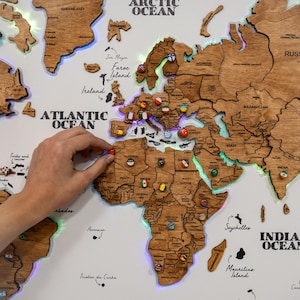 Op de afbeelding: Een houten wereldkaart met contouren van landen en vlagpinnen. De kaart wordt verlicht met kleurrijke LED-verlichting rond de continenten. De kaart omvat de Atlantische, Arctische en Indische Oceaan. Een hand plaatst een speld op de kaart.