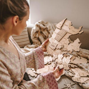 May include: A wooden map of North America, with the USA and Canada labeled. The map is made of light-colored wood and is being held by a person wearing a patterned robe. The map is detailed with state and province outlines.
