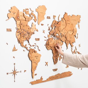 May include: A light brown wooden world map wall art featuring laser-cut continents, countries, and oceans. The map includes a compass rose, small wooden icons of ships and planes, and the text Antarctica. Numerous push pins mark locations.