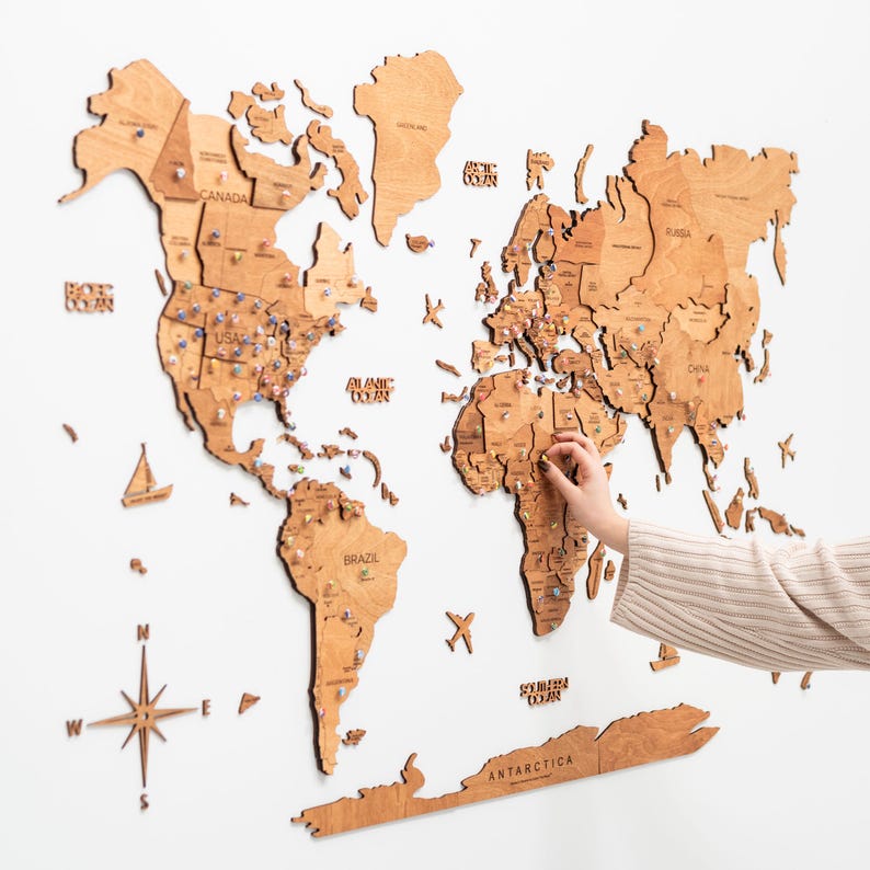 May include: A light brown wooden world map wall art featuring laser-cut continents, countries, and oceans. The map includes a compass rose, small wooden icons of ships and planes, and the text Antarctica. Numerous push pins mark locations.
