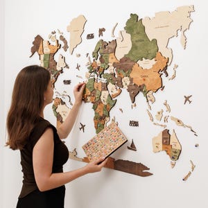 May include: A large wooden world map wall art piece featuring multi-colored continents in shades of green, brown, and natural wood. The map includes laser-cut country names, ocean labels, and small wooden airplane and sailboat icons.