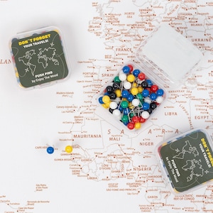 Peut inclure: Une boîte transparente remplie d'épingles à nourrice colorées, avec une carte en arrière-plan. La boîte porte l'inscription "DON'T FORGET YOUR TRAVELS!" et "PUSH PINS by Enjoy The Wood."