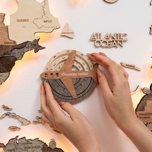 May include: A wooden world map with illuminated edges and individual country pieces. A wooden airplane-shaped travel counter with the text "Countries Visited" is being placed on the map. The counter has a dial with numbers and a compass design.