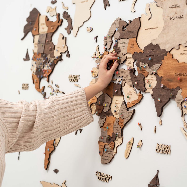 May include: A large wooden world map wall art piece featuring various shades of brown and beige wood tones. The map includes laser-cut country names, continents, and oceans, with small colorful push pins inserted into various locations on the map.