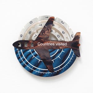 May include: Wooden travel-themed wall decor featuring a dark brown airplane shape with the text "Countries visited." The airplane is set against a blue and white circular background with a compass design. A silver-coloured gear-shaped ring surrounds the airplane, with numbers 0-9.