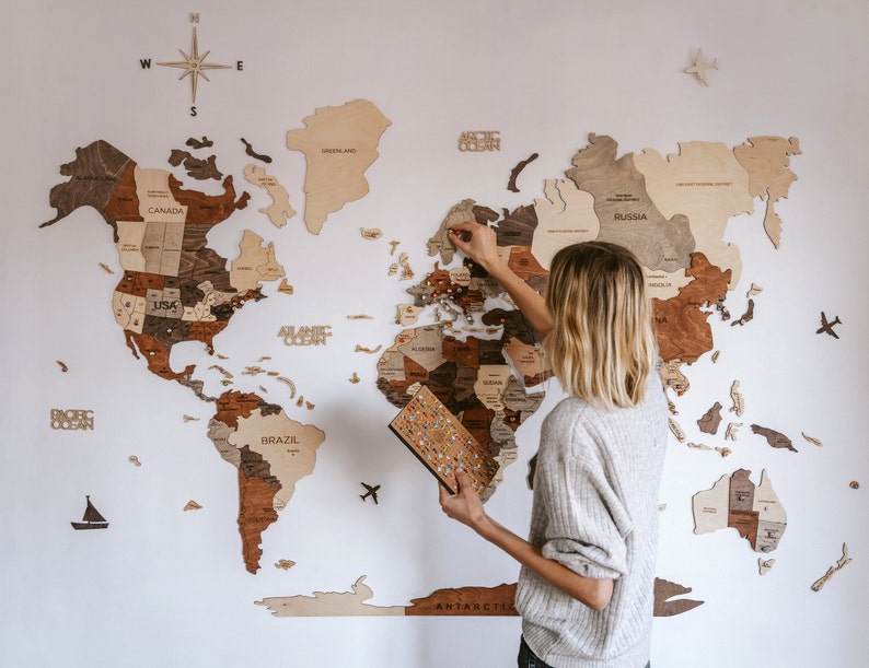 A woman with shoulder-length blonde hair, wearing a light grey knit sweater, stands in front of the map. She is facing the wall with her back to the camera, slightly turned, and appears to be placing a small flag pin into Norway. In her left hand, she holds a colorful flags pin set board, suggesting she is actively marking her travels or planning future adventures. This image depicts a cozy and creative moment centered around a large, beautifully crafted 3D wood world map mounted on white wall.