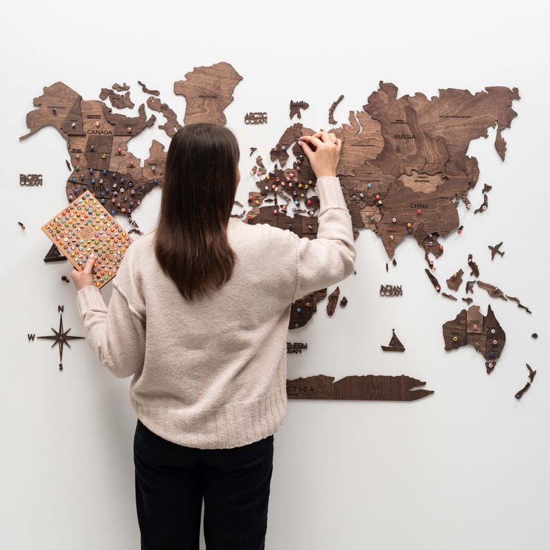 Puede incluir: Un mapa del mundo de madera con contornos de pa&iacute;ses y marcadores de alfiler est&aacute; montado en una pared blanca. Una persona est&aacute; colocando alfileres en el mapa. El mapa es de madera marr&oacute;n oscuro.