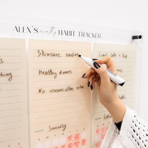 May include: A clear acrylic monthly habit tracker with a beige background and black text. The tracker is divided into sections for each day of the month, with space to write habits. The text "ALEX'S monthly HABIT TRACKER" is at the top. A hand is holding a white marker, writing on the tracker.