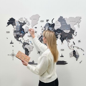 May include: A large, multi-piece wooden world map wall art installation featuring various shades of gray and black. The map includes labeled continents, countries, and oceans, with small decorative icons like airplanes and ships placed throughout.