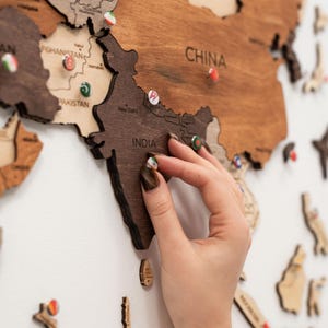 Puede incluir: Un mapa del mundo de madera con contornos de pa&iacute;ses y alfileres. El mapa est&aacute; hecho de madera marr&oacute;n oscura y clara. Una mano est&aacute; colocando un alfiler en India. Los alfileres tienen peque&ntilde;os dise&ntilde;os de banderas.