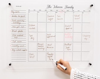 May include: Clear acrylic family calendar with the text "The Johnson Family" at the top. The calendar has a to-do list on the left and a monthly grid for scheduling appointments and events. A hand is writing on the calendar with a white marker.