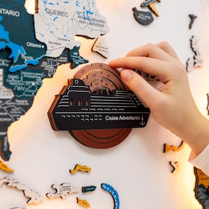 May include: A black and brown wooden cruise ship-shaped travel tracker with the words "Cruise Adventures" printed on it. The tracker is being placed on a white surface with a map of North America in the background. Other map pieces are scattered around.