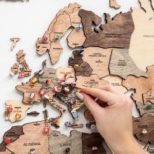 Puede incluir: Un mapa del mundo de madera con contornos de pa&iacute;ses y alfileres de bandera. El mapa est&aacute; hecho de madera marr&oacute;n clara y oscura. Una mano coloca un alfiler en el mapa, destacando un destino de viaje. El mapa incluye los nombres de pa&iacute;ses y regiones.