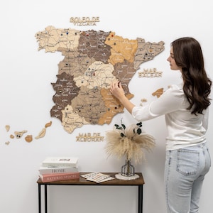 May include: A wooden map of Spain with various shades of brown and tan, mounted on a white wall. The map includes the names of cities and regions. A person is interacting with the map. Decorative elements include wooden lettering and small wooden islands.