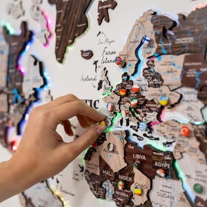 May include: A wooden world map with country outlines and pins. The map is lit with LED lights, and a hand is placing a pin on the map. The map includes country names and geographical features.