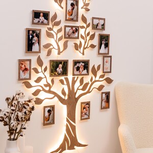 Puede incluir: Una decoraci&oacute;n de pared de madera con forma de &aacute;rbol con ramas y hojas, con luces enhebradas alrededor del tronco y las ramas. Hay 14 peque&ntilde;os marcos de fotos con fotos de personas en su interior, unidos al &aacute;rbol. El &aacute;rbol est&aacute; montado en una pared blanca.