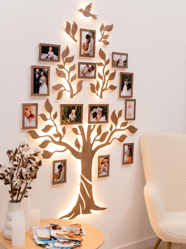 Puede incluir: Una decoraci&oacute;n de pared de madera con forma de &aacute;rbol con ramas y hojas, con luces enhebradas alrededor del tronco y las ramas. Hay 14 peque&ntilde;os marcos de fotos con fotos de personas en su interior, unidos al &aacute;rbol. El &aacute;rbol est&aacute; montado en una pared blanca.
