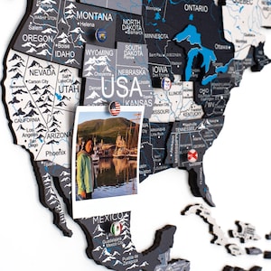 May include: A multi-layered wooden wall map of North America featuring black, white, and blue color schemes with printed state and country names. The map includes small circular pins with national flags and a photograph attached to the USA section.