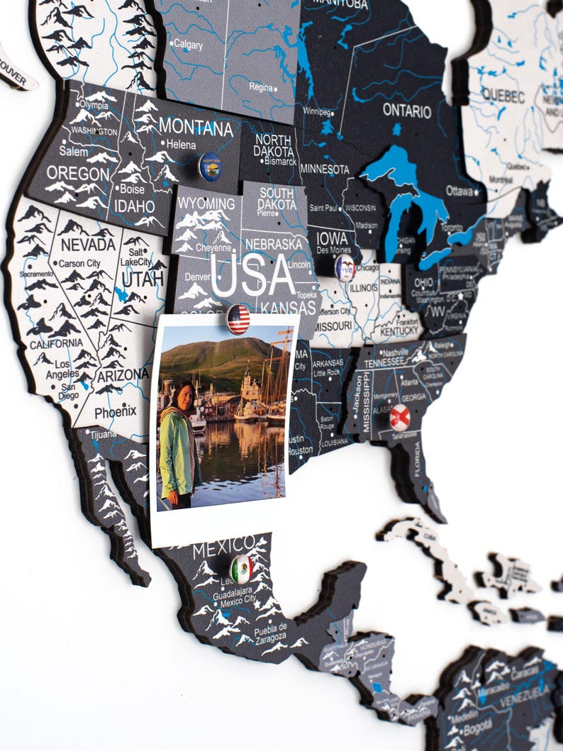 May include: A multi-layered wooden wall map of North America featuring black, white, and blue color schemes with printed state and country names. The map includes small circular pins with national flags and a photograph attached to the USA section.
