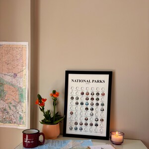 May include: A framed "National Parks" print with a black border, featuring illustrations of park icons. A small potted plant with orange flowers, a red mug, and a lit candle are on a desk. A map is visible on the wall.