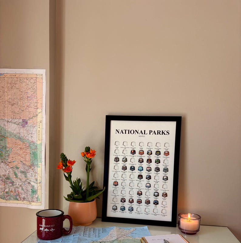 May include: A framed "National Parks" print with a black border, featuring illustrations of park icons. A small potted plant with orange flowers, a red mug, and a lit candle are on a desk. A map is visible on the wall.