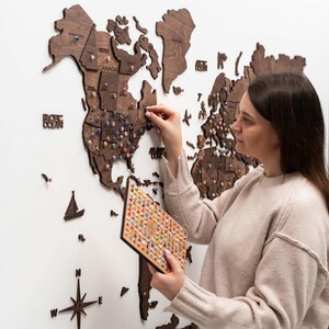 May include: A dark brown wooden world map wall art installation featuring laser-cut continents, countries, and states. The map includes decorative wooden icons like a sailboat and compass rose, with numerous colorful push pins inserted into the wood.