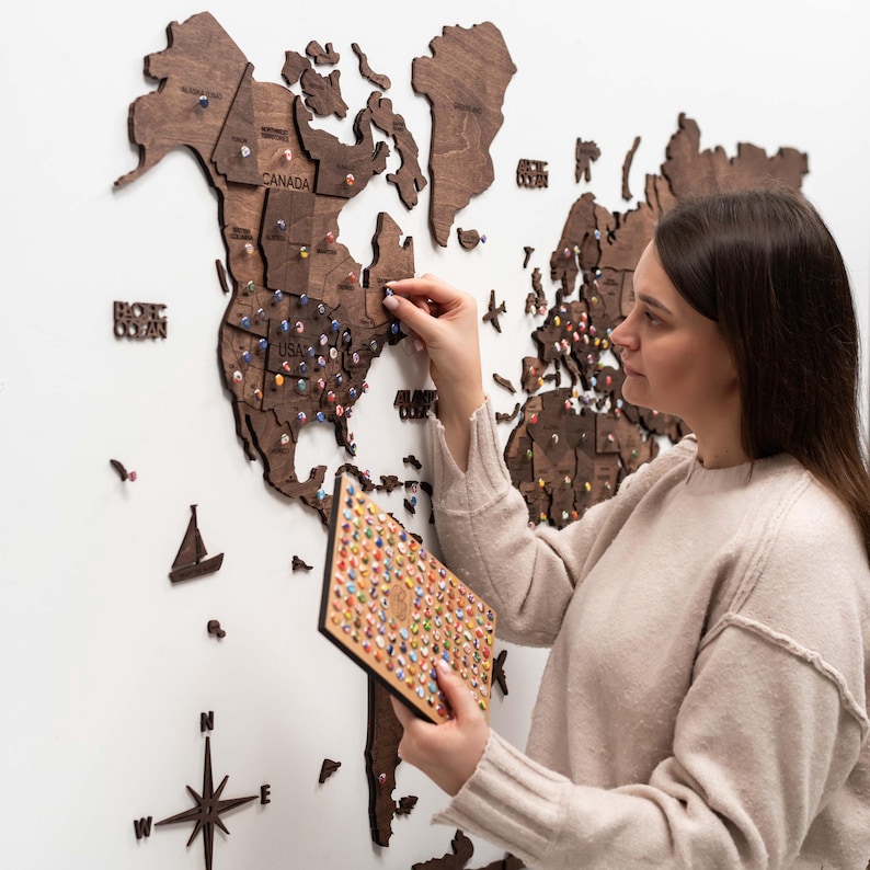 May include: A dark brown wooden world map wall art installation featuring laser-cut continents, countries, and states. The map includes decorative wooden icons like a sailboat and compass rose, with numerous colorful push pins inserted into the wood.