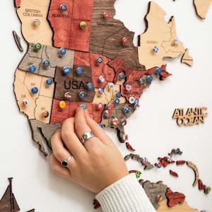 Puede incluir: Mapa del mundo de madera con contornos de estados y pa&iacute;ses. El mapa est&aacute; hecho de varios tonos de madera e incluye alfileres con banderas. El texto "USA" es visible. Las palabras "Atlantic Ocean" tambi&eacute;n son visibles.