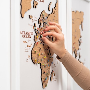Peut inclure: Une carte du monde en liège avec un cadre blanc, présentant les contours des pays et des épingles. La carte est étiquetée "ATLANTIC OCEAN" et comporte diverses épingles drapeau. Une main place une épingle sur la carte.