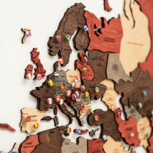 Può includere: Una mappa in legno dell'Europa con vari paesi in diverse tonalità di marrone e rosso. Piccoli segnaposti con bandiera contrassegnano le località. La mappa è montata su una superficie bianca.