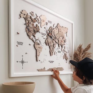 May include: A framed wooden world map with country outlines and pins. The map is mounted on a white wall and includes labels for oceans and a compass rose. The frame is white.