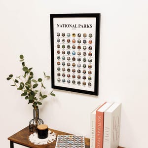May include: A framed print titled "NATIONAL PARKS VISITED" with a black frame. The print features a grid of colorful illustrations of park badges. The print is hanging on a white wall above a wooden table with a vase of greenery, a candle, and books.