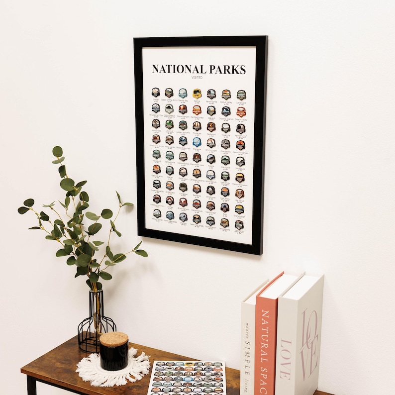 May include: A framed print titled "NATIONAL PARKS VISITED" with a black frame. The print features a grid of colorful illustrations of park badges. The print is hanging on a white wall above a wooden table with a vase of greenery, a candle, and books.