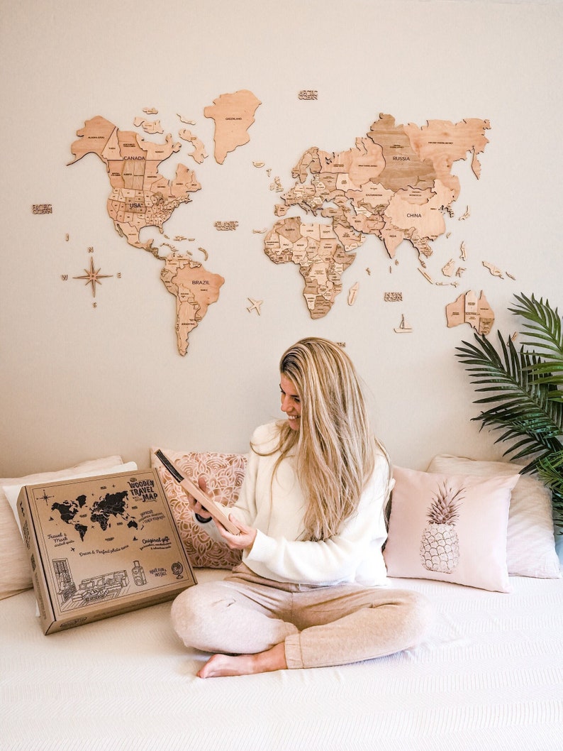 May include: A wooden world map decorates a wall, with a woman holding a wooden travel map. A box with the words "Wooden Travel Map" is on the bed. The map is a light brown color, and the box is brown. Pink pillows and a plant are also visible.