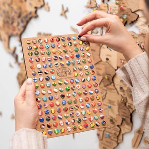 May include: A close-up of a wooden world map with colourful push pins representing different countries. The map is being held by a person's hand. The push pins are arranged in a grid pattern and feature flags from around the world. The text "Enjoy the Wood" is printed on the map.