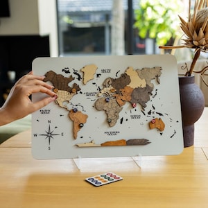 Peut inclure: Un panneau rectangulaire de couleur claire présentant une carte du monde en bois superposé avec diverses nuances de brun et de beige. La carte inclut des étiquettes pour les océans et une rose des vents. Des marqueurs circulaires sont posés.