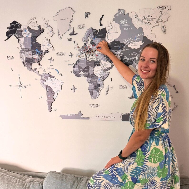 May include: A large, multi-piece wooden world map wall art in shades of gray, black, and white. The map features detailed country names, borders, and decorative icons like airplanes and ships. It is mounted on a wall, displaying global geography.