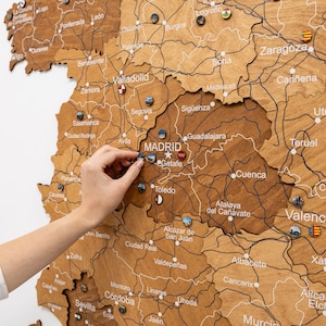 Peut inclure: Carte en bois de l'Espagne avec les noms des villes et les routes grav&eacute;s. De petites &eacute;pingles color&eacute;es marquent les lieux. Une main place une &eacute;pingle pr&egrave;s de Madrid. La carte est de couleur marron clair.