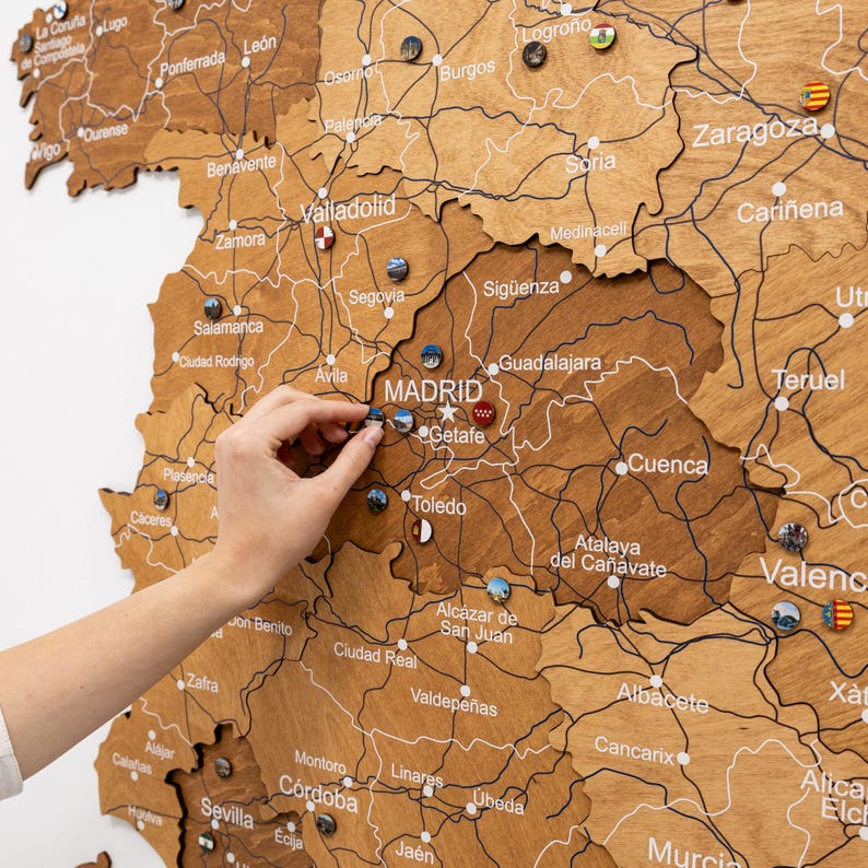 Peut inclure: Carte en bois de l'Espagne avec les noms des villes et les routes grav&eacute;s. De petites &eacute;pingles color&eacute;es marquent les lieux. Une main place une &eacute;pingle pr&egrave;s de Madrid. La carte est de couleur marron clair.
