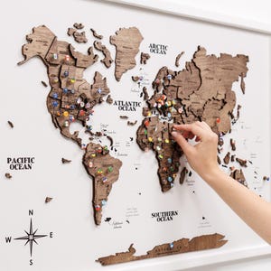 May include: A wooden world map with country outlines and colourful flag pins. The map is mounted on a white background and framed in white. The oceans are labelled. A hand is placing a pin on the map.