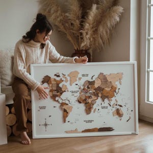 May include: A framed wooden world map with a white border. The map features various shades of brown and tan to represent different countries and continents. The oceans are white, and the map includes compass rose details.