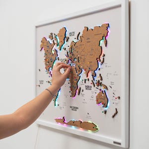 May include: A framed world map with a white border, featuring a cork map with colorful push pins. The map is illuminated with a multi-colored LED light strip around the edges. A hand is placing a pin on the map.