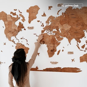 Pu&ograve; includere: Una grande mappa del mondo in legno con contorni dettagliati dei paesi, etichettati con i nomi dei paesi. La mappa &egrave; realizzata in legno marrone chiaro ed &egrave; montata su una parete bianca. Il testo "XL prime Oak" si trova nell'angolo in alto a destra. Una persona indica la mappa.