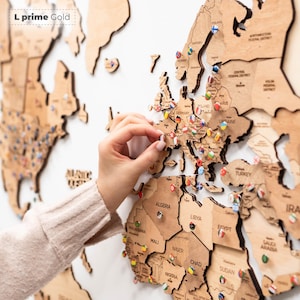 Pu&ograve; includere: Una mappa del mondo in legno con contorni di paesi e spilli. La mappa &egrave; realizzata in legno marrone chiaro ed &egrave; montata su una superficie bianca. Gli spilli sono colorati e rappresentano diversi paesi. Il testo "L prime Gold" &egrave; in alto a sinistra.